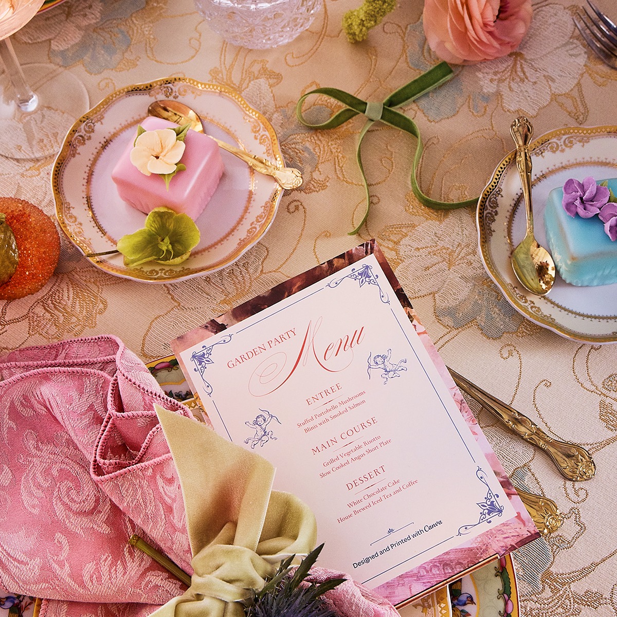 Close-up of a party invitation designed on Canva sits on a plate. In the background is a rococo party tablescape with colorful flowers, plates and treats that dot the table. 