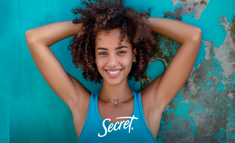 Mulher jovem com cabelos cacheados e sorriso radiante, vestindo uma blusa de alça azul, levantando os braços acima da cabeça. Ela está apoiada contra uma parede texturizada em tons de azul e verde turquesa, com o logo da Secret visível em sua blusa.